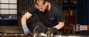 A technician wearing safety glasses and gloves inspects a large metal shaft using a measuring tool.